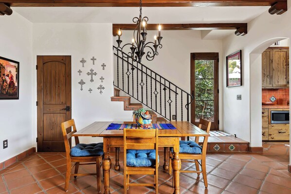 Un comedor con una mesa y sillas de madera, una escalera con barandilla de hierro forjado y una cocina visible a través de un arco.