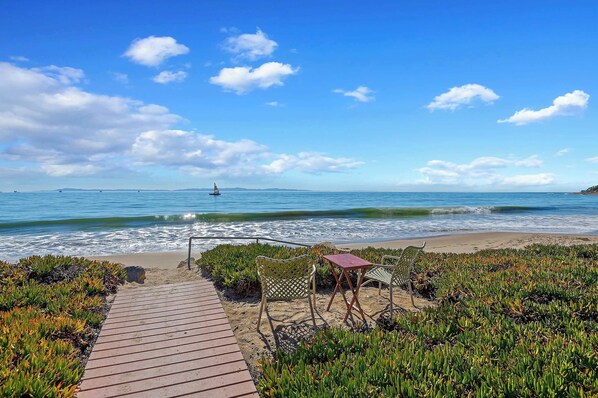 A beach with a wooden walkway leading to a seating area with chairs and a table.
