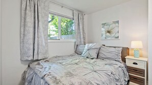 Een slaapkamer met een bed, gordijnen, een raam, een nachtkastje met een lamp en een kunstwerk aan de muur.