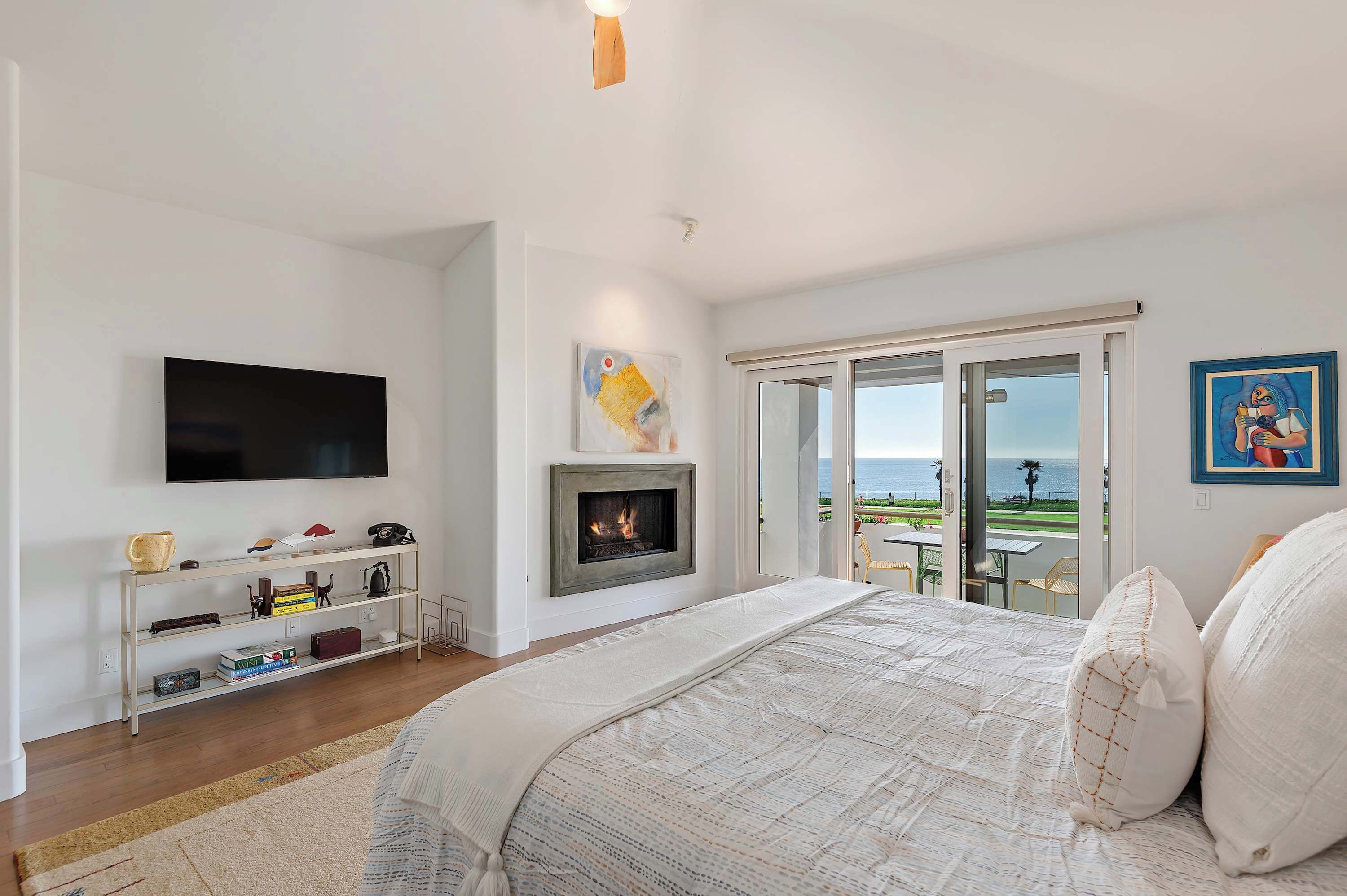 A bedroom with a large bed, a fireplace, a TV, and a balcony with a view of the sea.