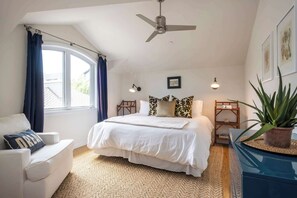 Et soveværelse med en stor seng, ventilator i loftet, et vindue med gardiner, et sengebord med lampe og en potteplante.