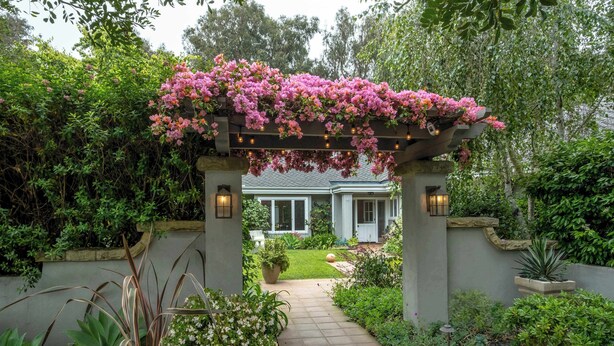 Un sentier de jardin menant à une maison surmontée d’une pergola recouverte de fleurs roses.