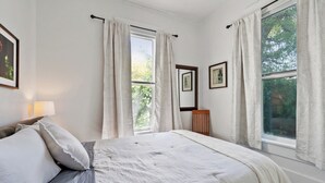 Un dormitorio con cama, dos ventanas con cortinas, una mesita de noche con lámpara y cuadros enmarcados en la pared.