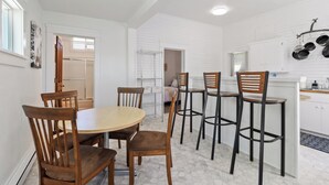 Un comedor con una mesa redonda y sillas de madera, una cocina con armarios blancos y ollas colgando, y un pasillo con una puerta que da a otra habitación.