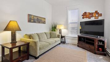 Una sala de estar con un sofá, dos mesitas auxiliares, una lámpara, un televisor y un elemento decorativo en la pared.
