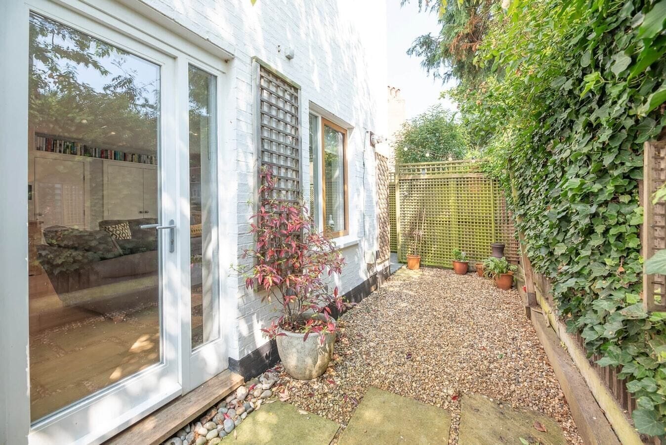 Une maison blanche avec une terrasse en gravier, un pot de fleurs et une clôture en bois recouverte de lierre.