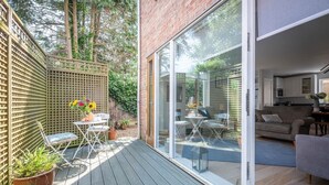 A modern patio with a wooden deck, a glass door leading to a living area, and a small table with chairs. - Skimming Stones, Southwold (Suffolk)