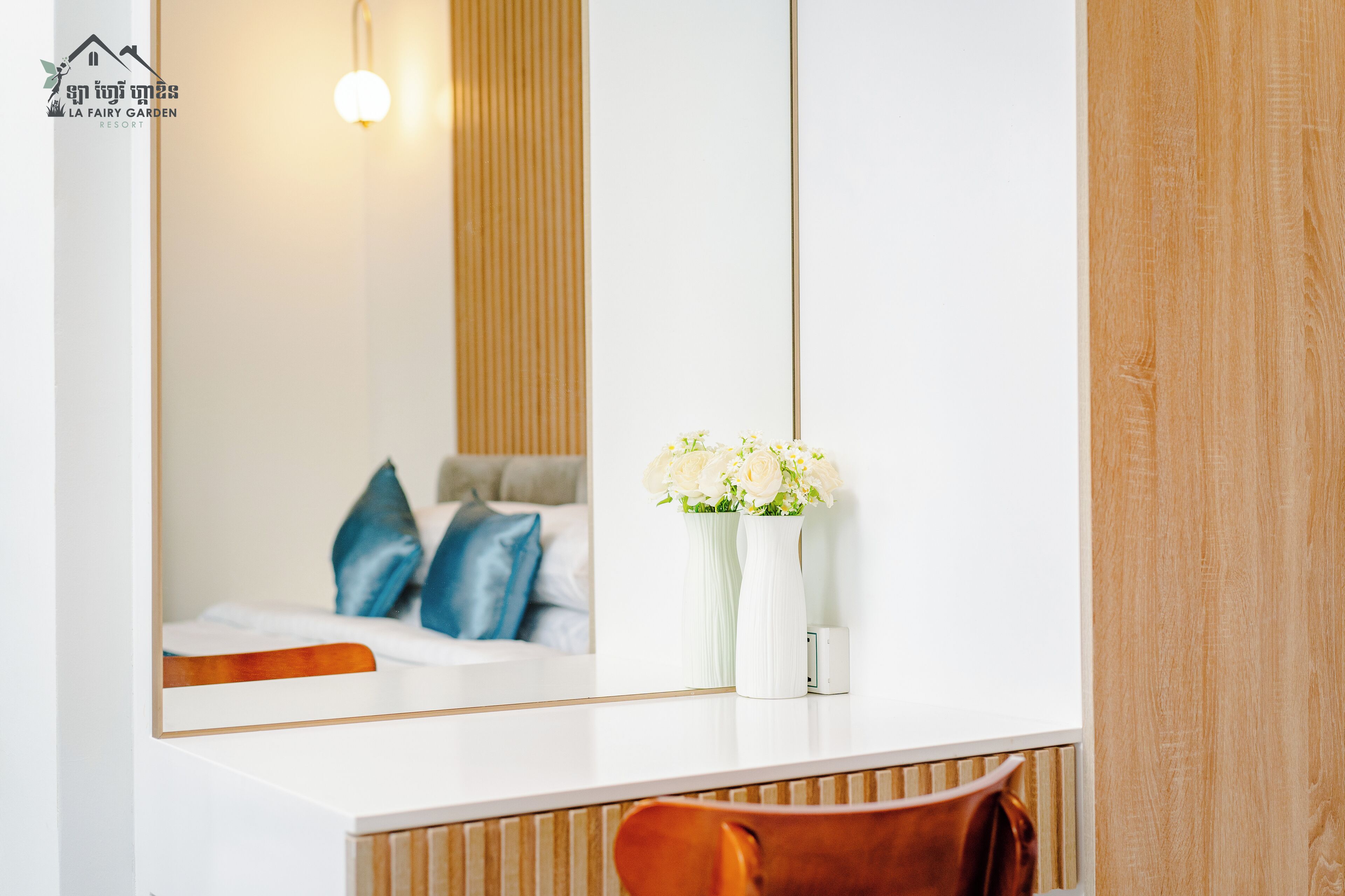 A modern hotel room with a large mirror, a wooden accent wall, a white bedside table with two vases of flowers, a brown chair, and a bed with blue pillows.