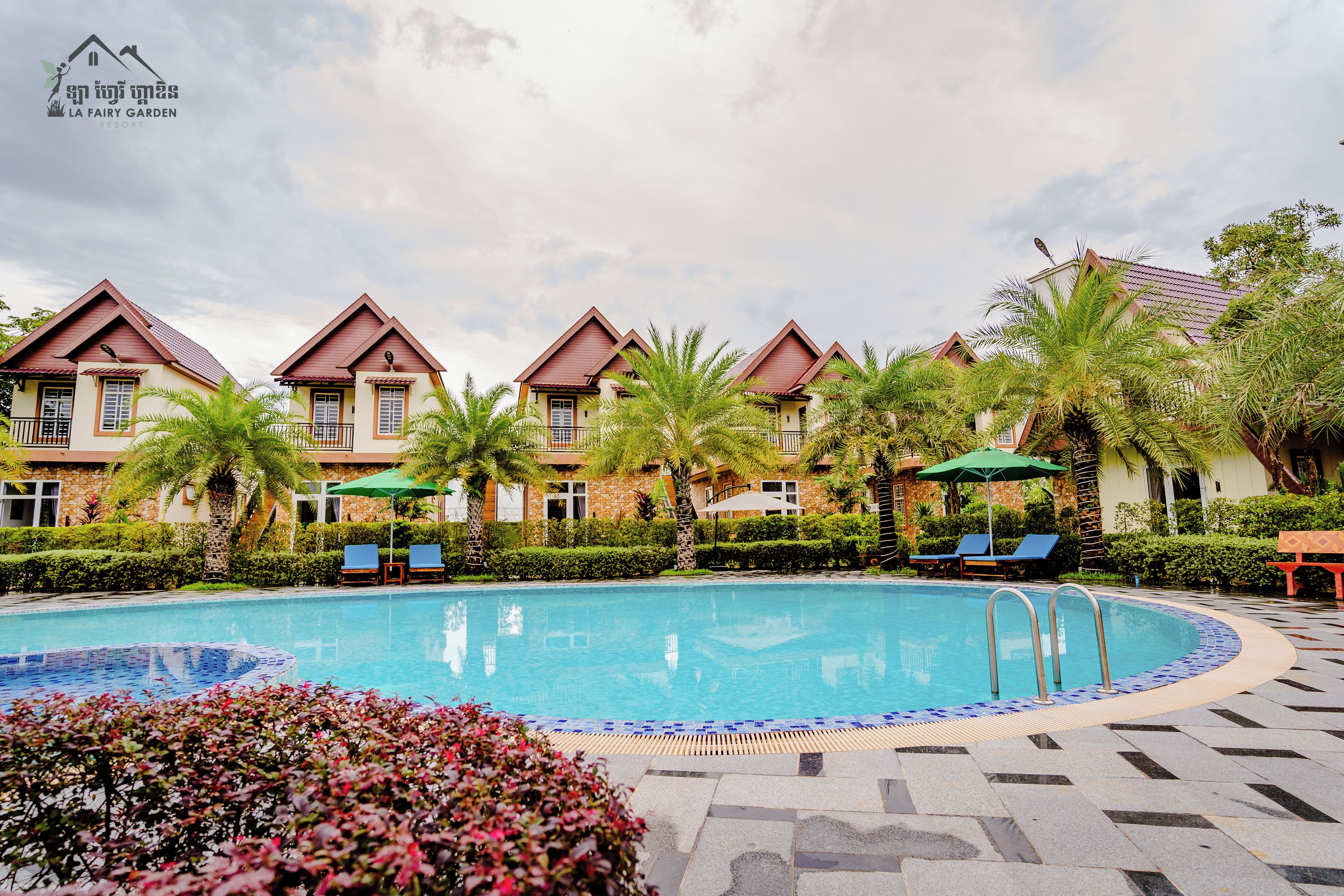 A swimming pool with lounge chairs and umbrellas, surrounded by palm trees and a building with a red roof.