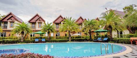 A swimming pool with lounge chairs and umbrellas, surrounded by palm trees and a building with a red roof.