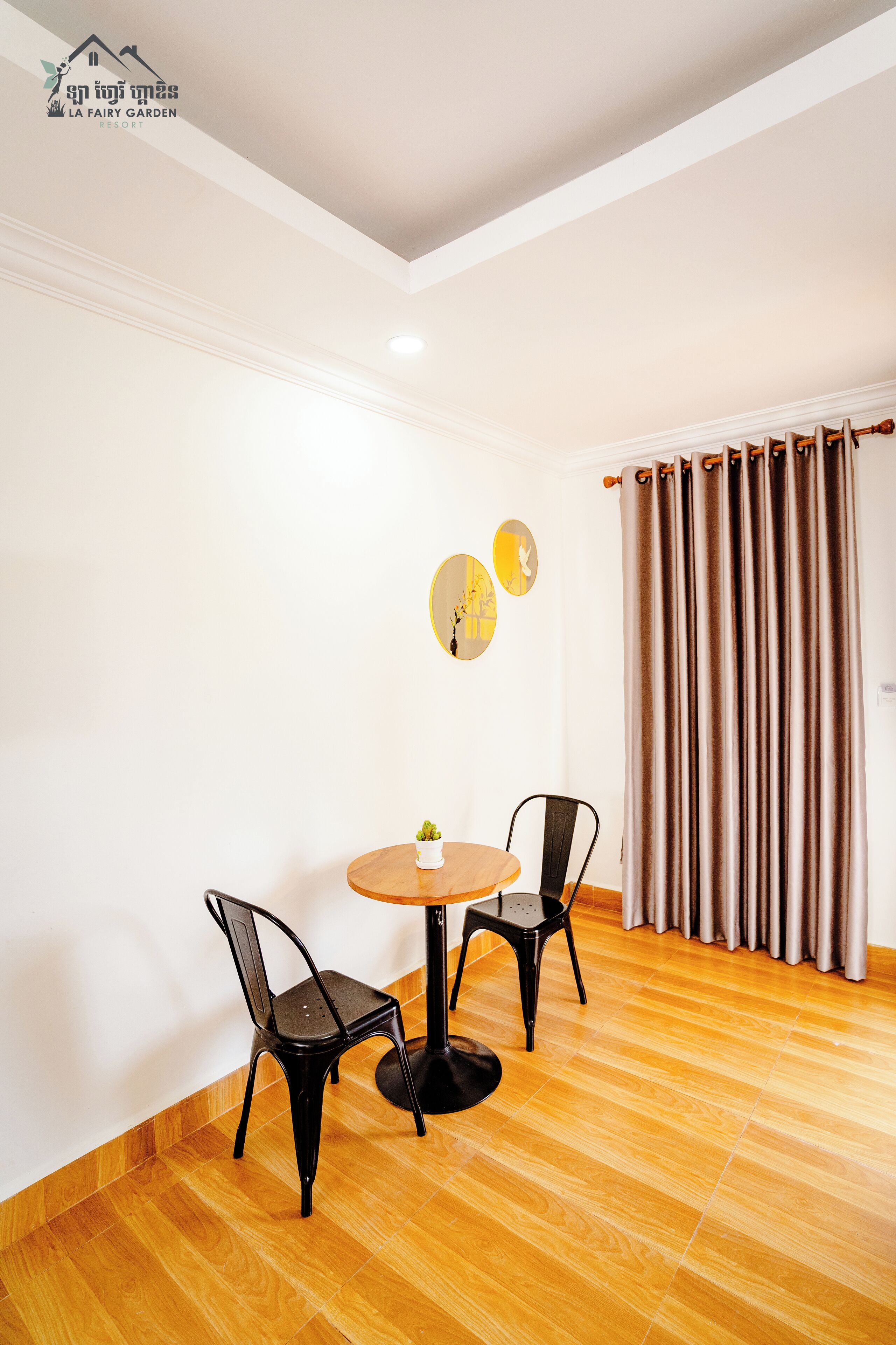 Una habitación con una mesa redonda de madera, dos sillas negras, una pequeña planta en maceta y una cortina.