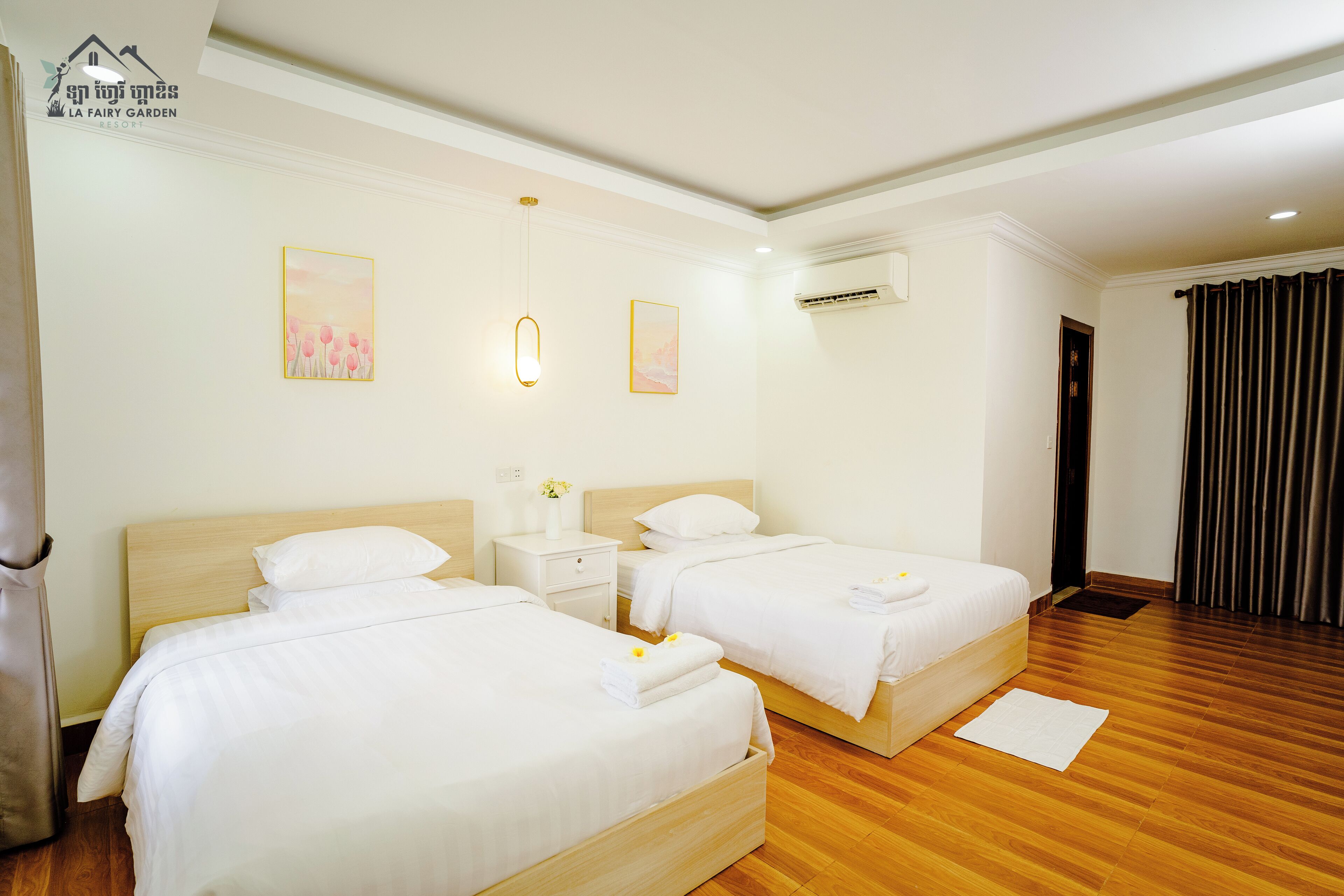 A hotel room with two beds, a wooden floor, a white wall, and a ceiling light.