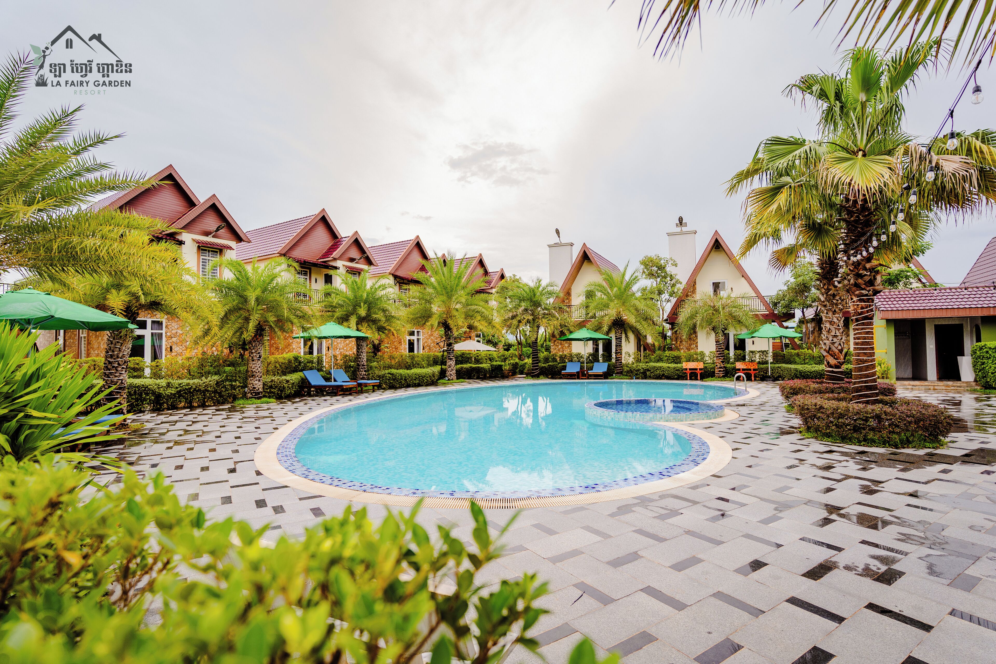 A pool area with a tiled deck, palm trees, and a building with a red roof.