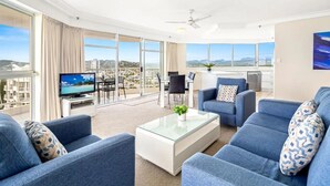 Ein geräumiges Wohnzimmer mit großem Fernseher, bequemen Sitzmöglichkeiten und einem Balkon mit Blick auf die Stadt.