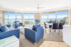 A modern living room with blue sofas, a glass coffee table, and a dining area with a white table and dark chairs. Large windows offer a view of the ocean. - Oceania Oceanview 2 bed Unit (Gold Coast)