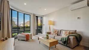 A modern living room with a sofa, ottoman, coffee table, and a lamp. There is a large window with curtains, an air conditioning unit, and a view of the outdoors. - The Beacon - Town Centre Apartment 620 (Queenstown)