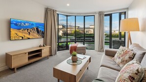 A modern living room with a large window, a flat-screen TV, a sofa, a coffee table, and a wooden TV stand. - The Beacon - Town Centre Apartment 620 (Queenstown)