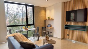 A modern living room with a sofa, a dining table, and chairs. There is a TV mounted on the wall and a window with a view of trees and buildings. - Cozy Aparta-Suite Studio | Rosa Negra (Medellín)
