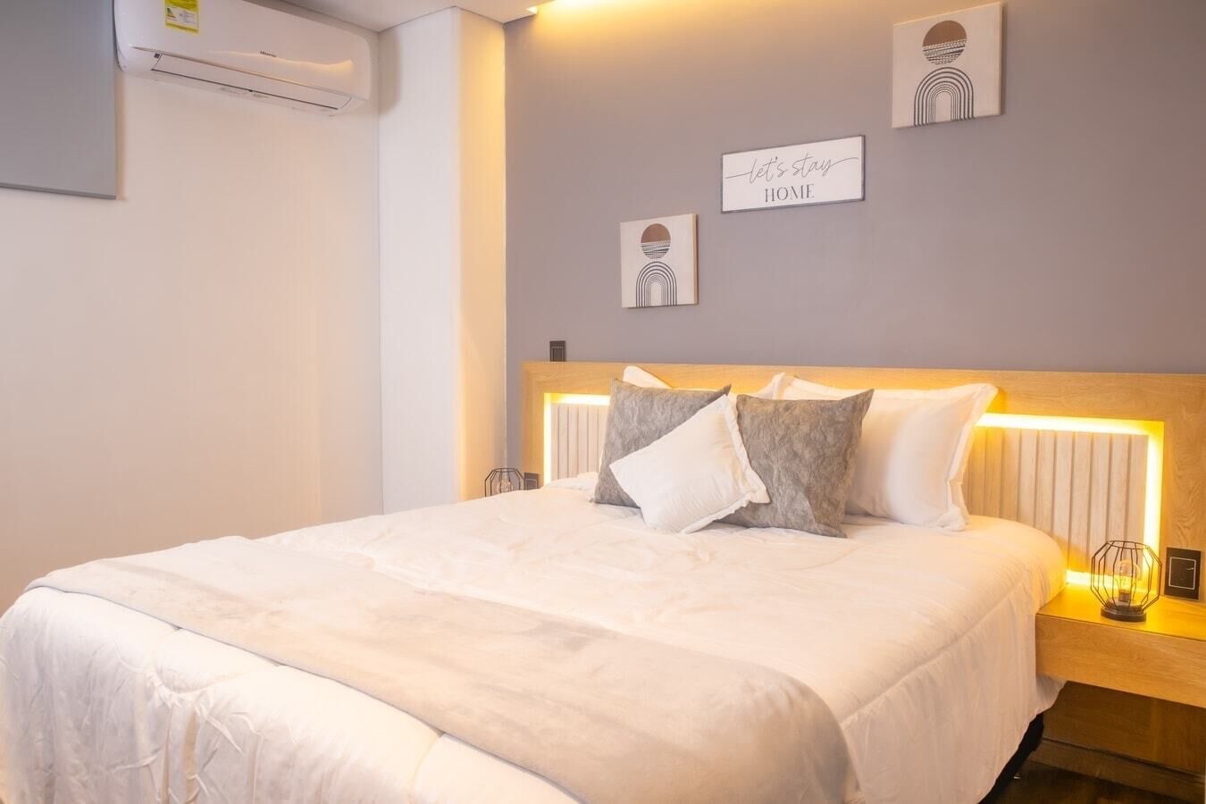 A neatly made bed with a white comforter and pillows, a headboard with integrated lighting, and a wall-mounted air conditioner.