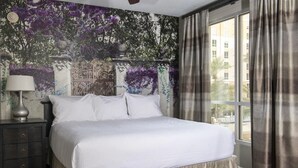 Un cuarto de hotel con una cama grande, una mesita de noche con lámpara, una ventana con cortinas y un mural en la pared.
