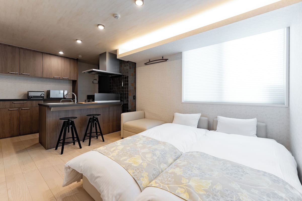 A modern bedroom with a large bed, a small kitchenette, and a window with blinds.