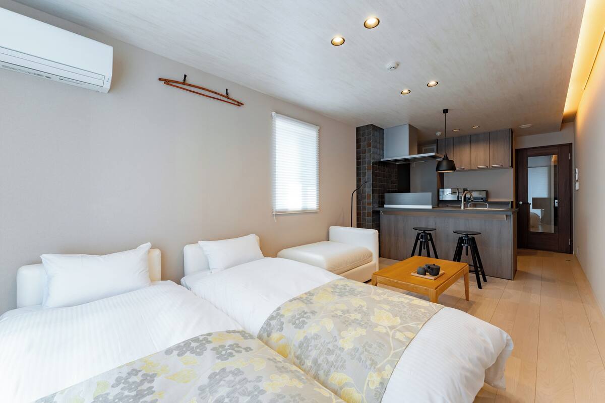 A modern hotel room with a large bed, a sofa, a small kitchen area, and a dining table.