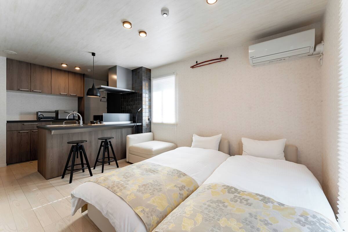 A modern hotel room with a kitchenette, a bed with floral bedding, a small seating area, and a window with blinds.