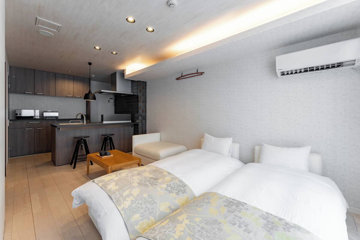 A modern hotel room with a large bed, a small desk, a kitchenette, and an air conditioning unit.