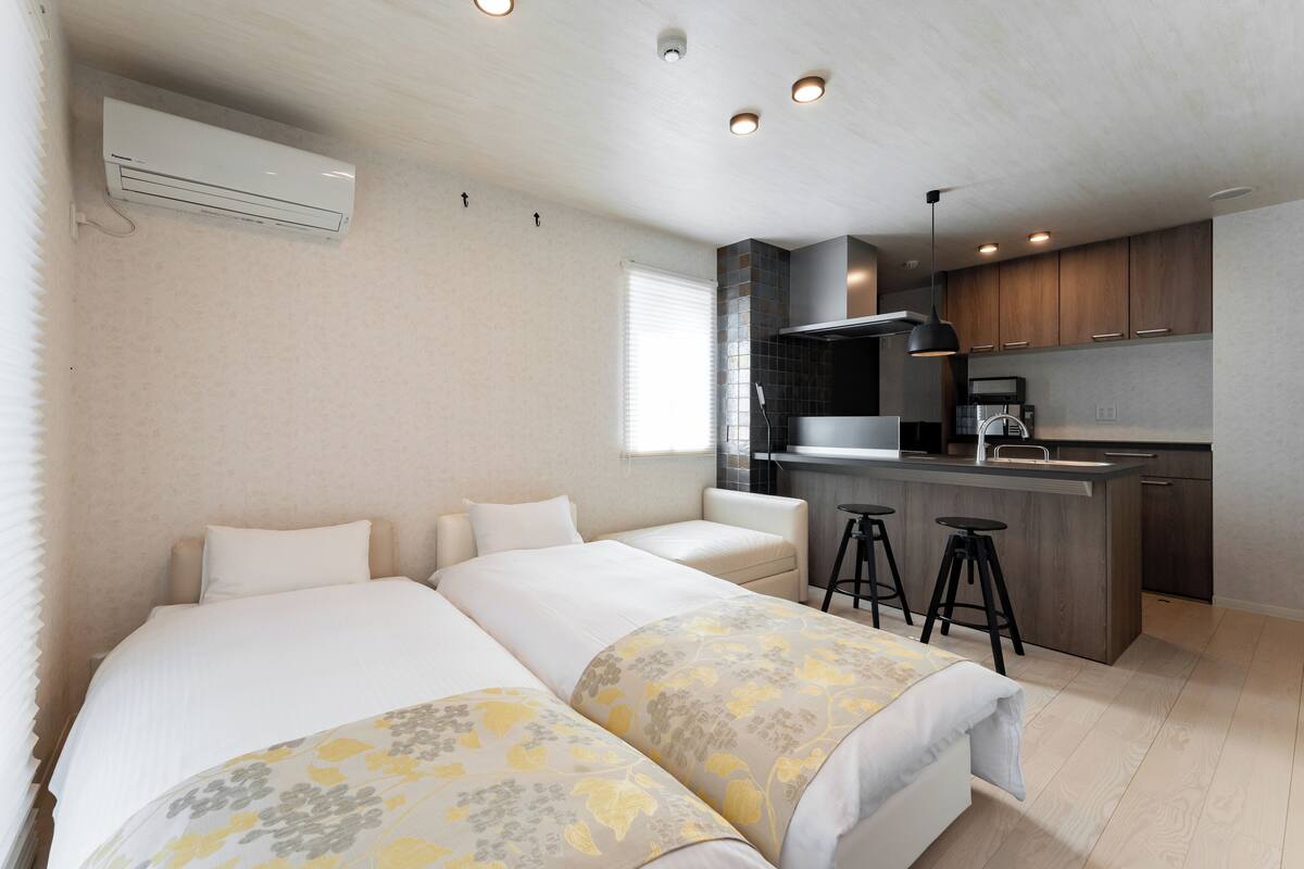 A modern hotel room with two beds, a kitchenette, and a bathroom.