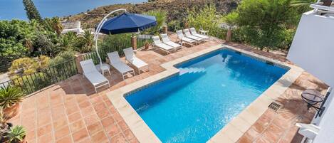 A rectangular swimming pool with a blue border, surrounded by white lounge chairs and a blue umbrella.