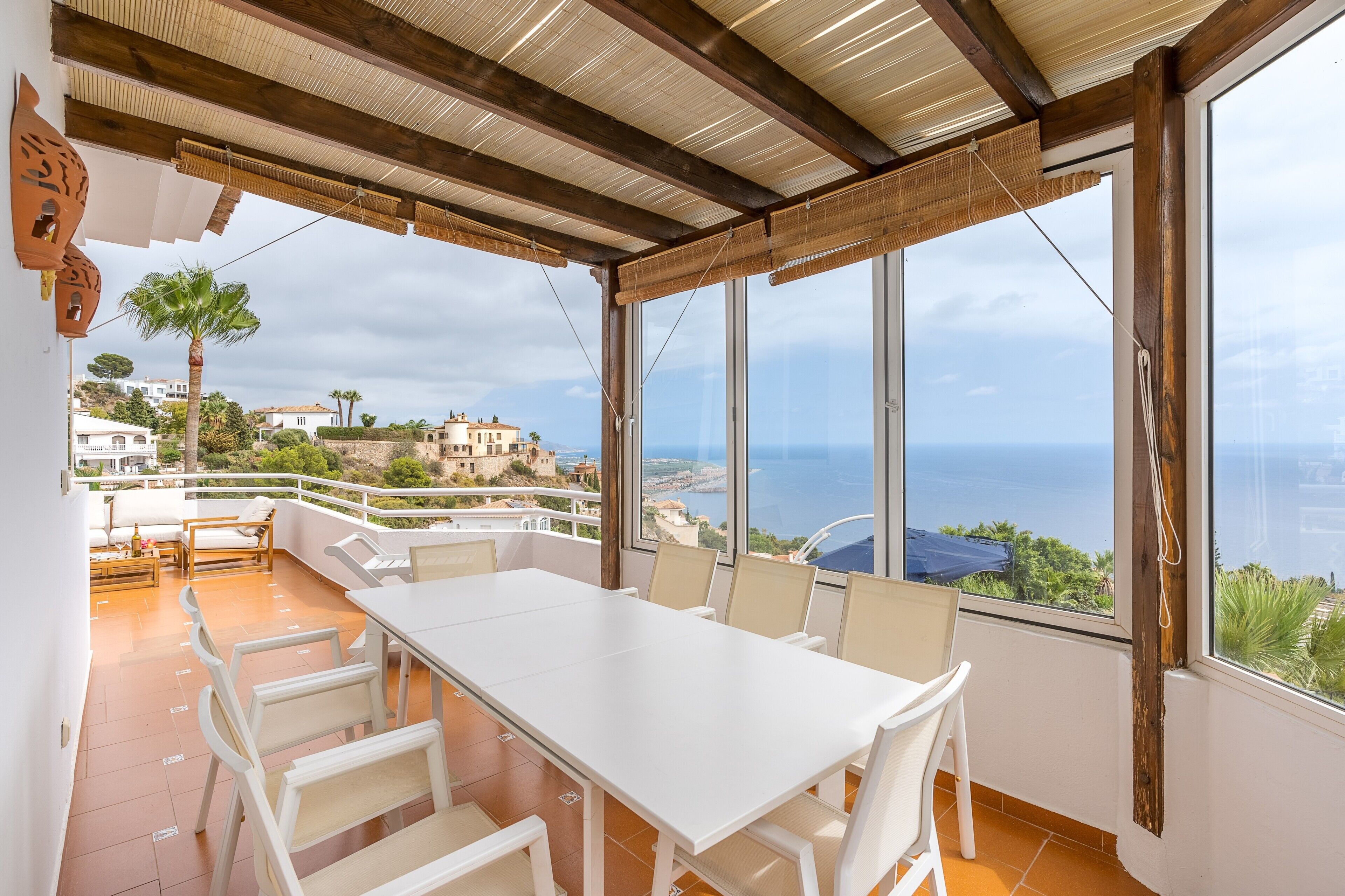 Eine Terrasse mit weißem Tisch und Stühlen, große Fenster mit Blick auf Meer und Gebäude sowie eine Holzbalkendecke.