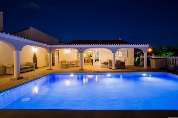 Une piscine avec une terrasse carrelée, un espace de détente couvert avec des chaises, et un bâtiment doté d’un toit en tuiles.