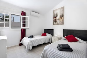 Une chambre d’hôtel avec deux lits, une fenêtre avec des rideaux, un climatiseur, une table de chevet et une commode, ainsi qu’un tableau accroché au mur.