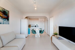 Un salon moderne avec un canapé, un coin repas comprenant une table et des chaises, une cuisine en arrière-plan et un téléviseur posé sur un meuble.