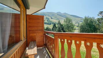 Un balcon doté d’une balustrade en bois, d’un banc et offrant une vue sur des collines verdoyantes et des montagnes.