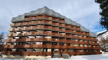 Un hôtel à plusieurs étages, doté de balcons, dont le sol est recouvert de neige, et qui se trouve au pied d’une chaîne de montagnes.