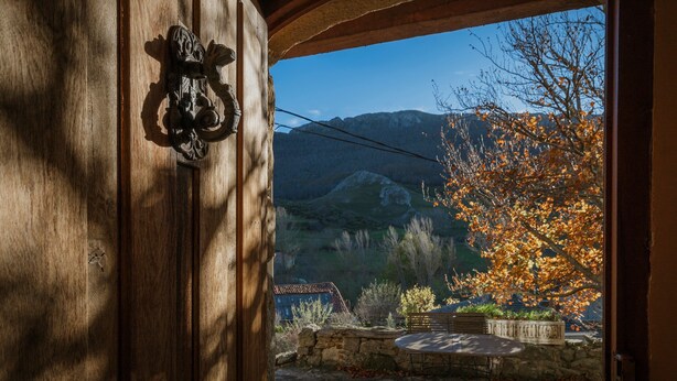 Una porta de fusta amb un mànec metàl·lic, una paret de pedra i una vista d'un paisatge de muntanya amb fullatge d'octubre.