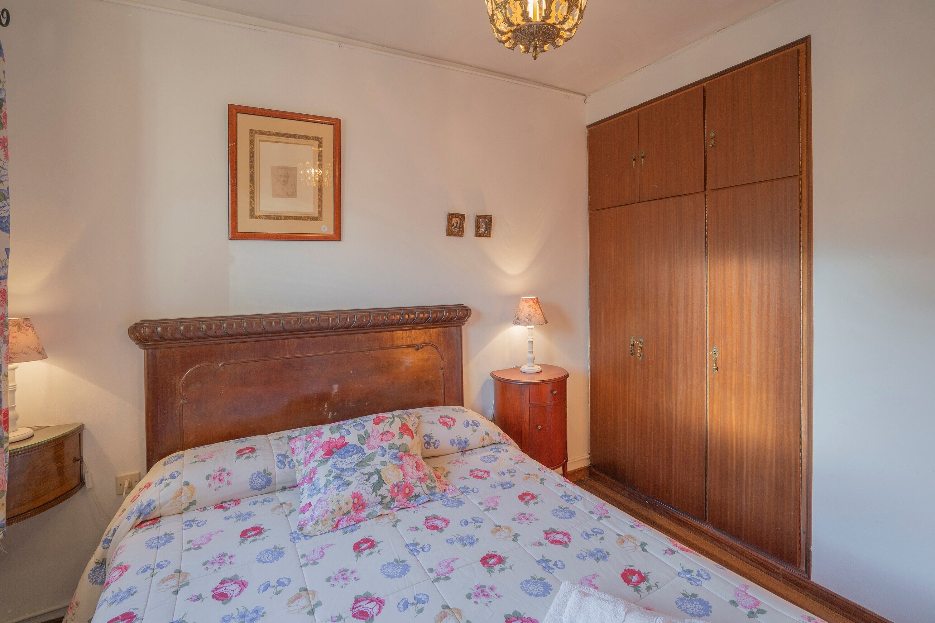 Een slaapkamer met een houten bed, een nachtkastje, een lamp, een kledingkast en een lijst met een afbeelding aan de muur.