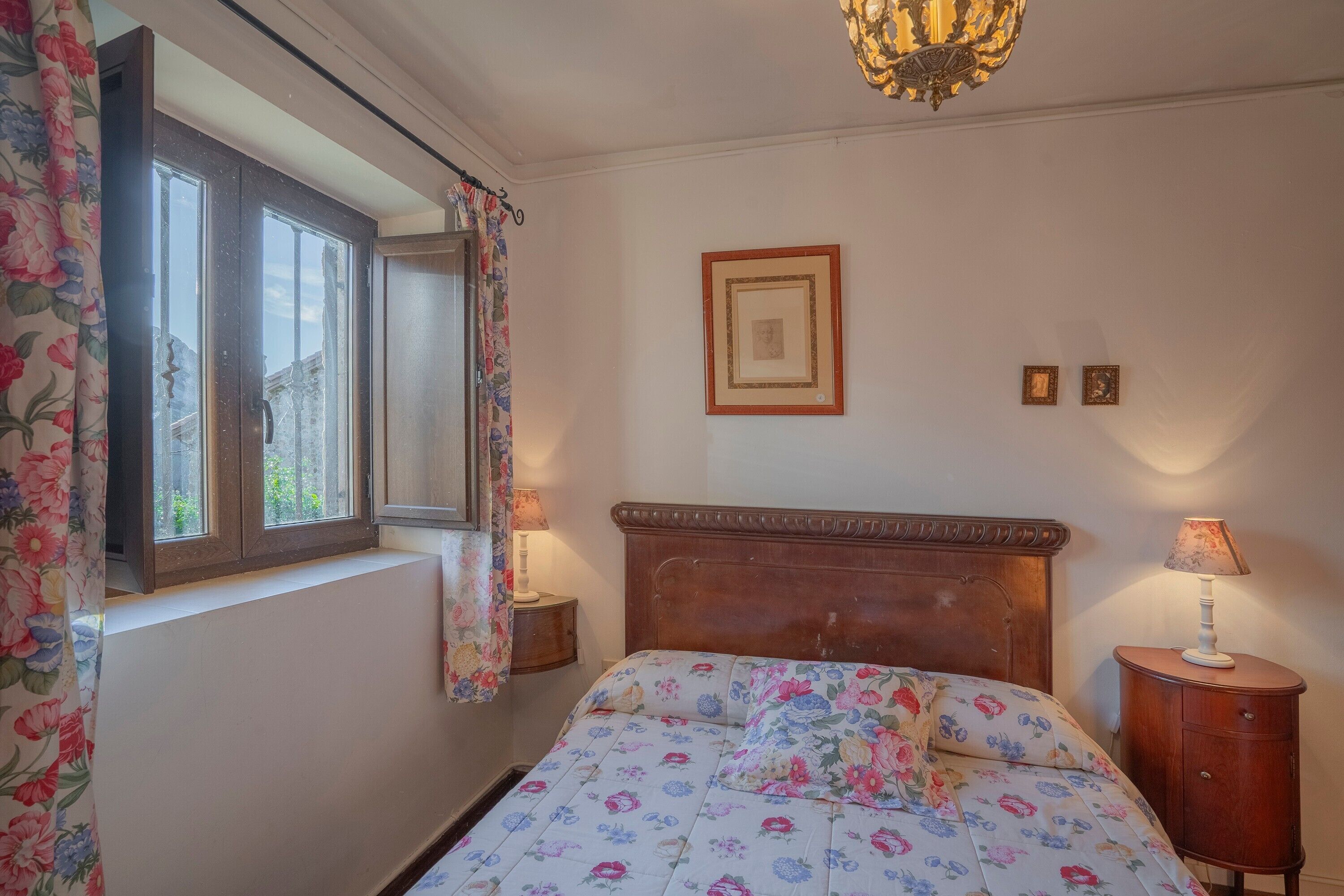 A bedroom with a bed, a window with curtains, a bedside lamp, and a framed picture on the wall.