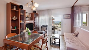 A living room with a glass dining table, wooden bookshelves, a sofa, and a view of the sea through a window.