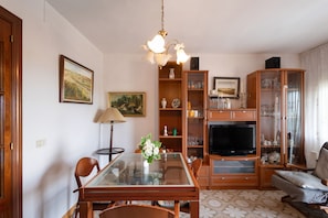 Ein Wohnzimmer mit einem Esstisch aus Glas, Holzstühlen, einem Fernseher und einer Vitrine mit Glastüren.