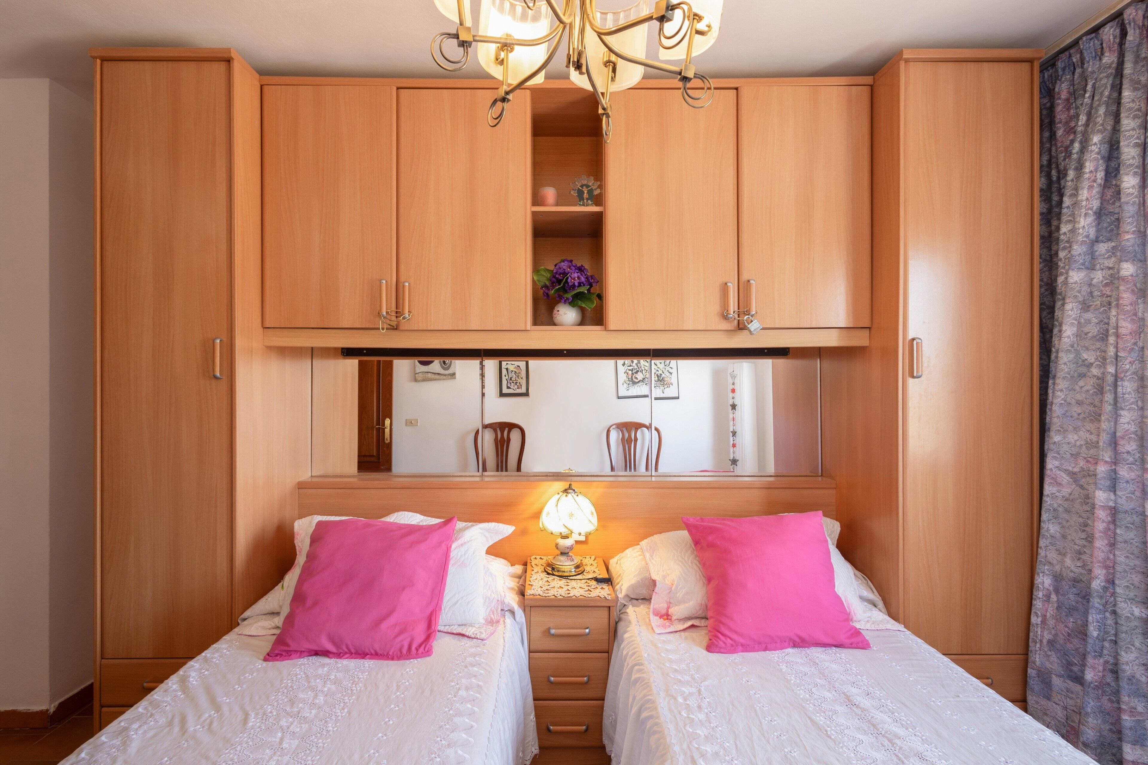 Un dormitorio con dos camas, una mesita de noche con lámpara, un armario de madera y un espejo sobre las camas.