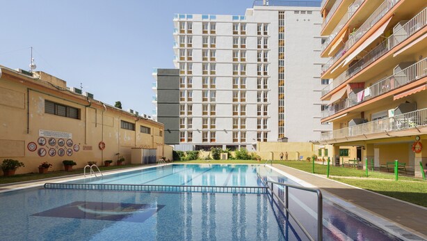 A swimming pool with a clear blue water surface, surrounded by a low fence. In the background, there is a tall residential building with multiple balconies and windows.