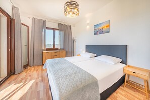 A bedroom with a bed, a wooden nightstand, a window with curtains, a painting on the wall, and a ceiling light fixture. - Melissa 1 - paradise views of the sea and mountains, large garden (Korfu)