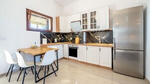 A modern kitchen with white cabinets, a stainless steel refrigerator, a wooden dining table with chairs, and a black marble backsplash. - Melissa apartment - stunning sea views, large garden and parking (Korfu)