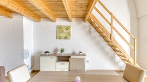 Un interior moderno con una escalera de madera, una zona de comedor con mesa y sillas, un escritorio con plantas y una pared con un cuadro.
