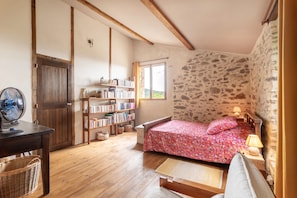 Une chambre avec un plancher en bois, un lit recouvert d’une couvre-lit à motifs floraux, une bibliothèque, un ventilateur et une fenêtre donnant sur un beau paysage.