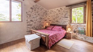 Een slaapkamer met een stenen muur, een houten bedframe, een nachtkastje met een lamp, een rieten mand en uitzicht op bomen door een raam.