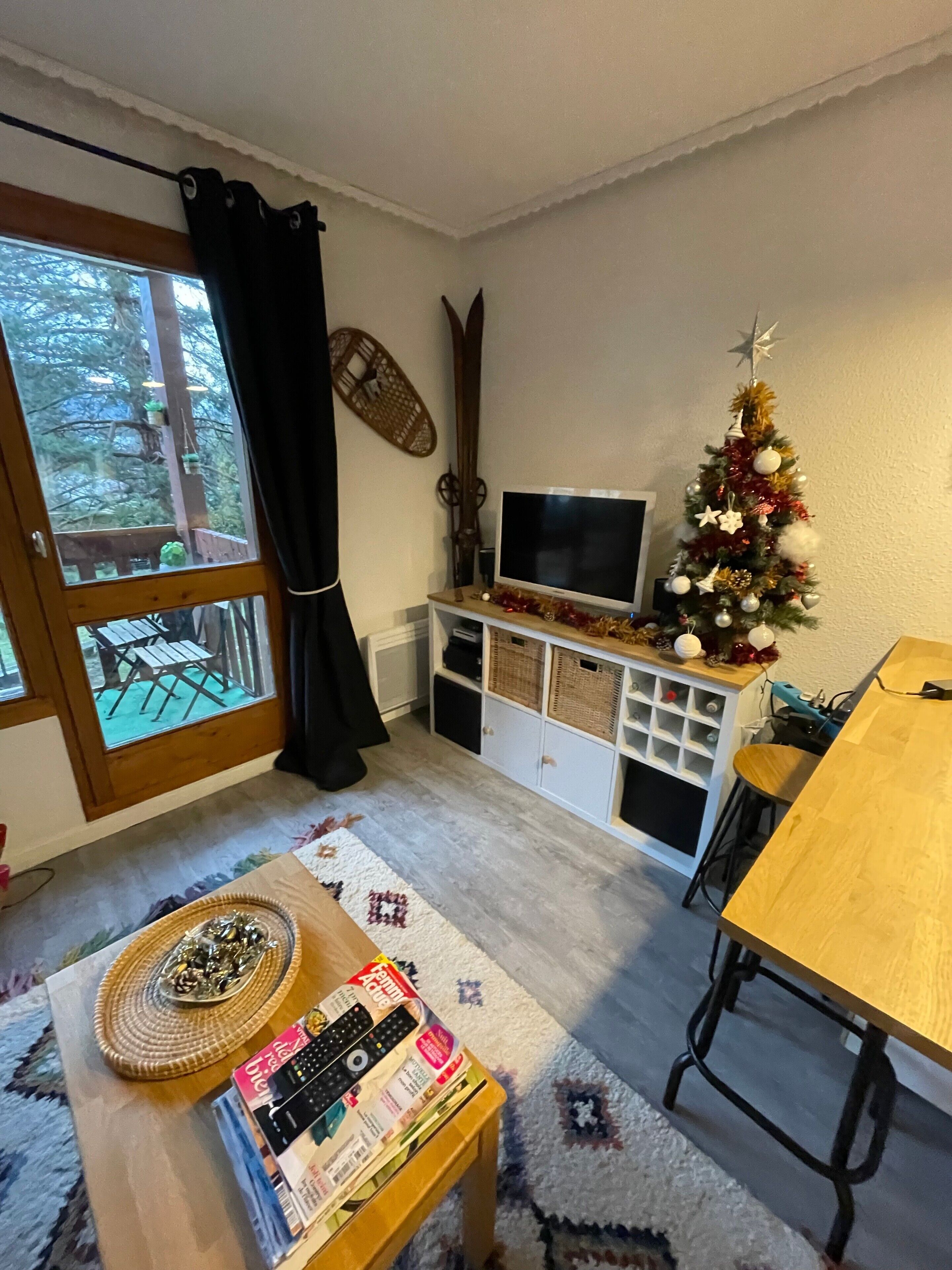 A living room with a TV, a Christmas tree, a wooden table, and a window with a view of trees.