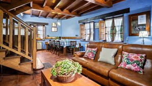 Una escalera de madera conduce a un comedor con una mesa y sillas, un sofá de cuero con cojines florales, y un cesto de flores sobre una mesa.